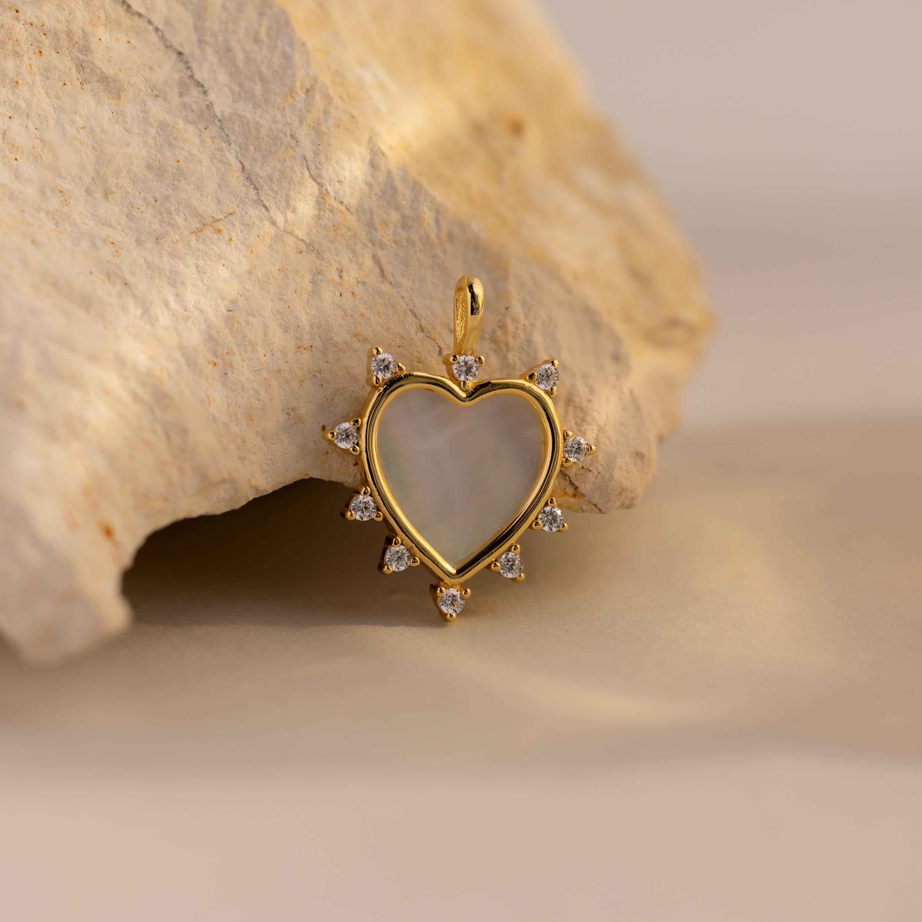 A single gold Starburst Pearl Heart Charm is displayed leaning against a beige stone, glowing under natural light.
