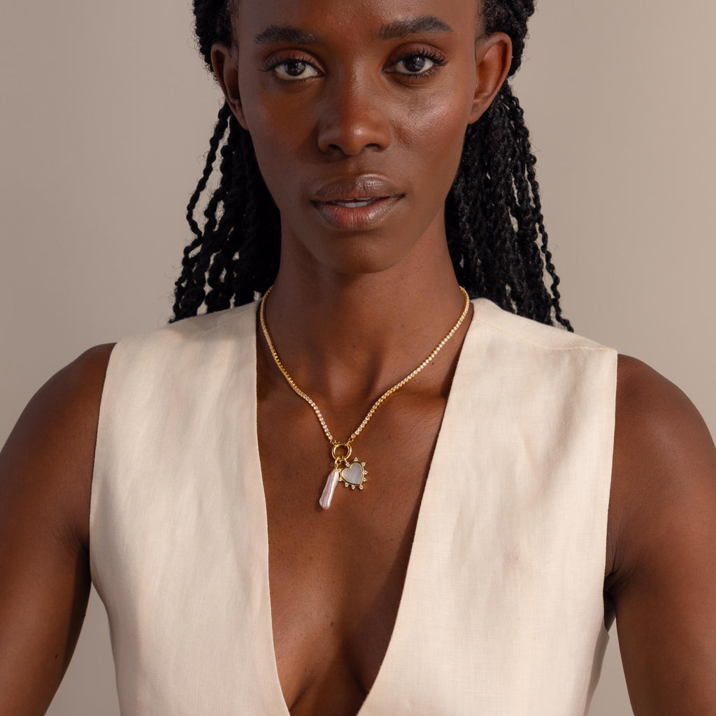 A model wears the gold Starburst Pearl Heart Charm on a diamond tennis rolo necklace with pearl charm, paired with a minimalist cream outfit.