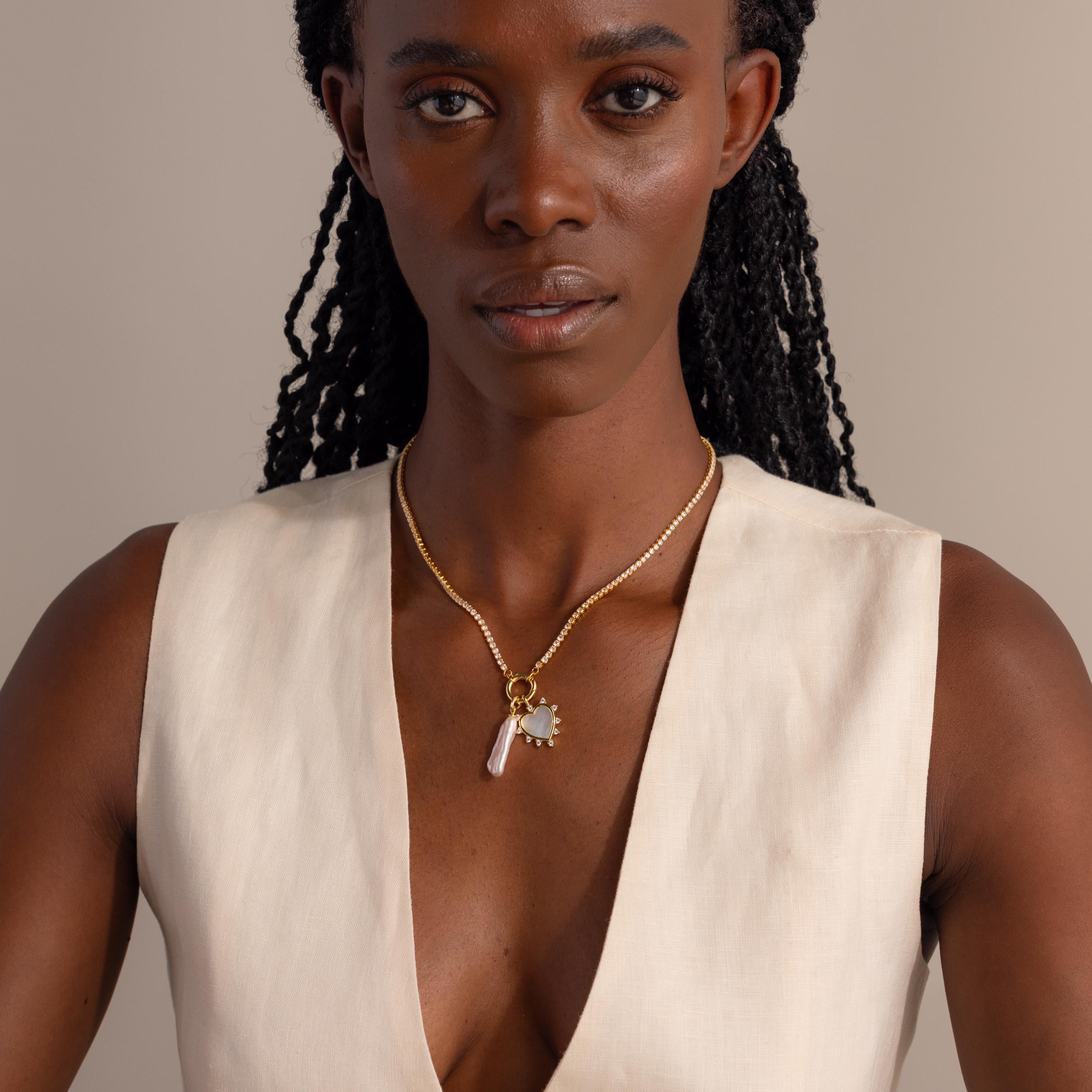 A model wears the gold Starburst Pearl Heart Charm on a diamond tennis rolo necklace with pearl charm, paired with a minimalist cream outfit.