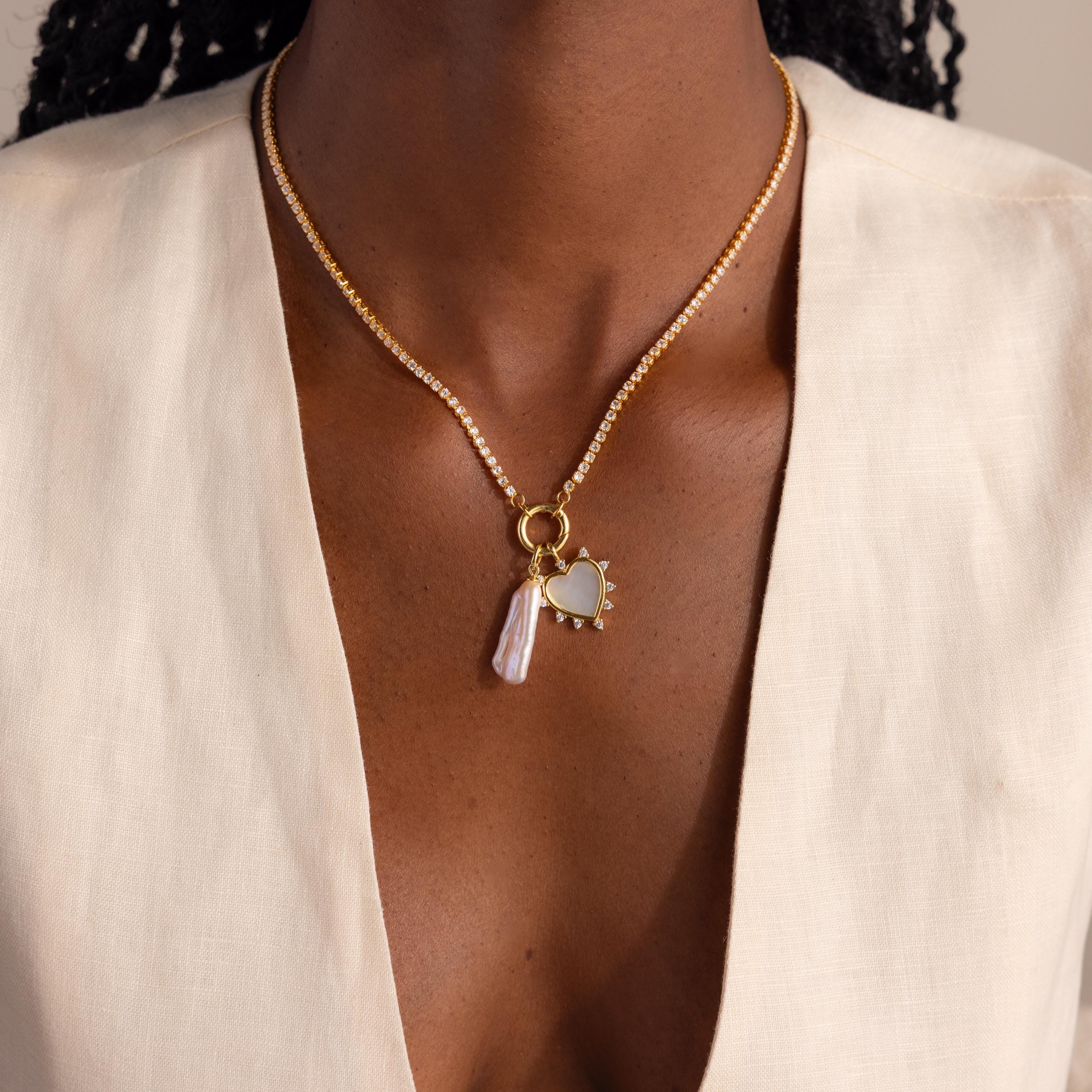 A close-up of a gold diamond tennis necklace worn around the neck, featuring the Starburst Pearl Heart Charm and a vertical pearl charm.
