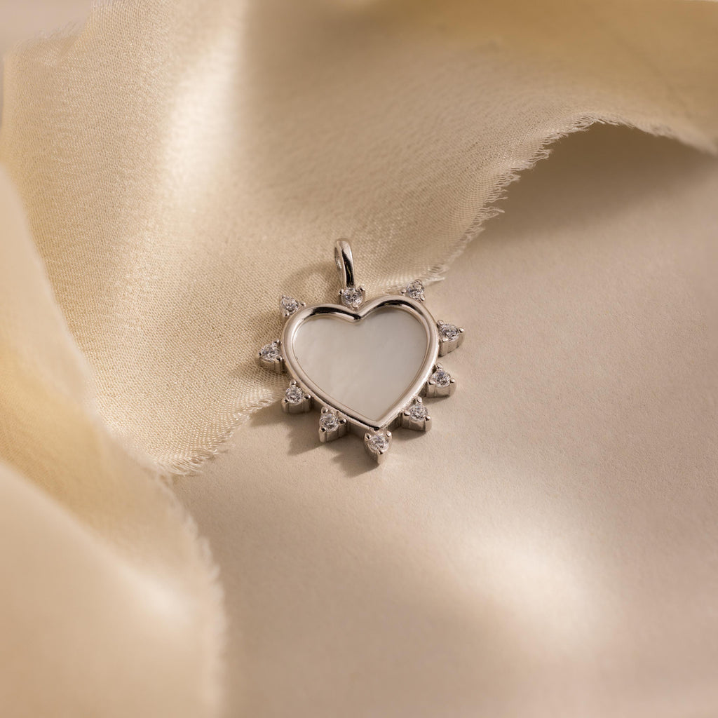 The silver version of the Starburst Pearl Heart Charm lies on a flowing, light satin fabric with its cubic zirconia accents catching soft light.