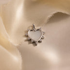 The silver version of the Starburst Pearl Heart Charm lies on a flowing, light satin fabric with its cubic zirconia accents catching soft light.