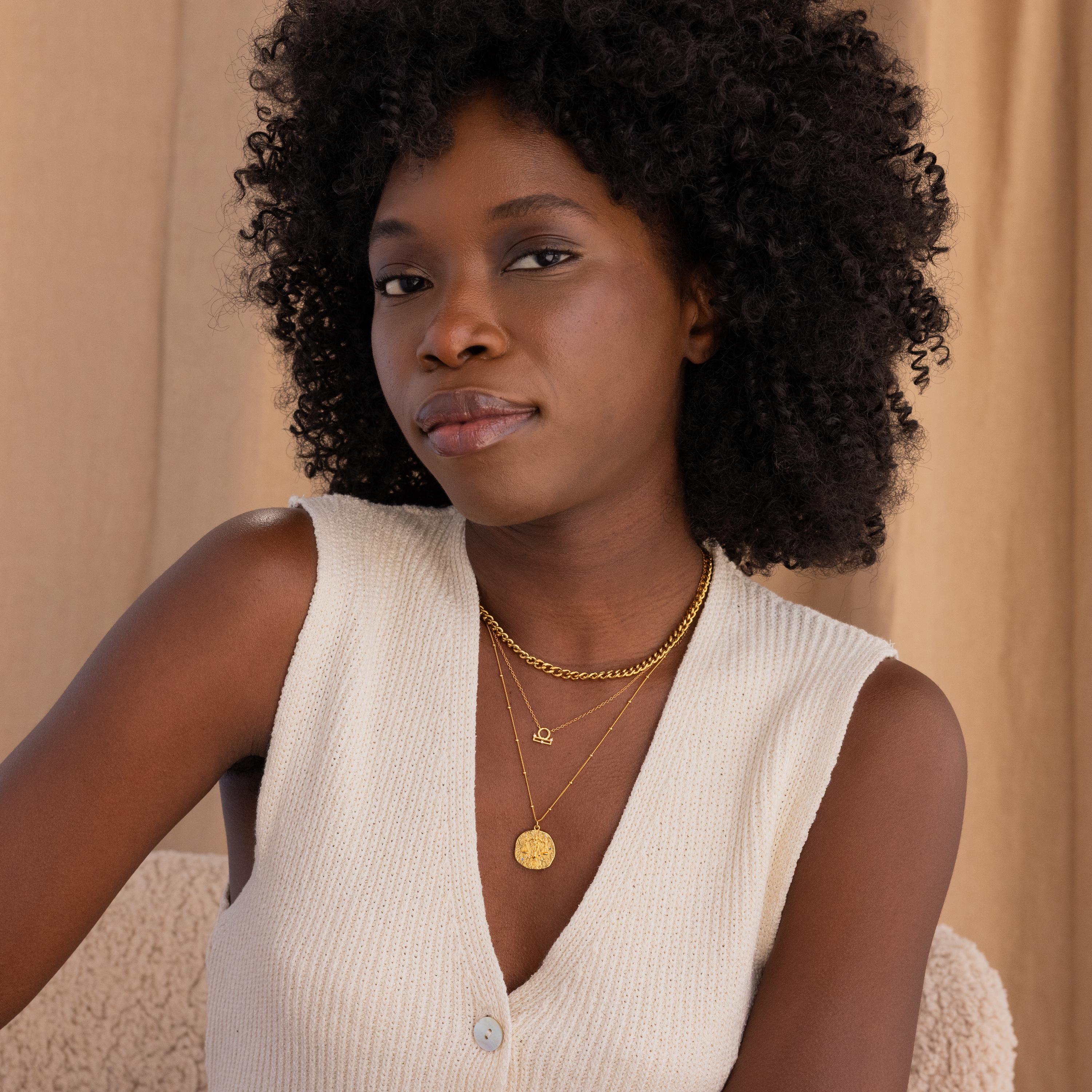 Wide view shot of model posing, wearing a gold libra zodiac necklace, layered with a thick gold curb chain on top and a hammered zodiac circle pendant on bottom.