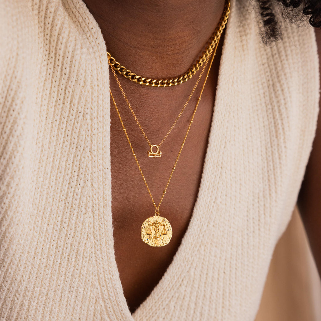 Model wears a gold CZ diamond libra zodiac charm necklace, layered with a thick gold curb chain on top and a hammered libra zodiac circle coin pendant on bottom.