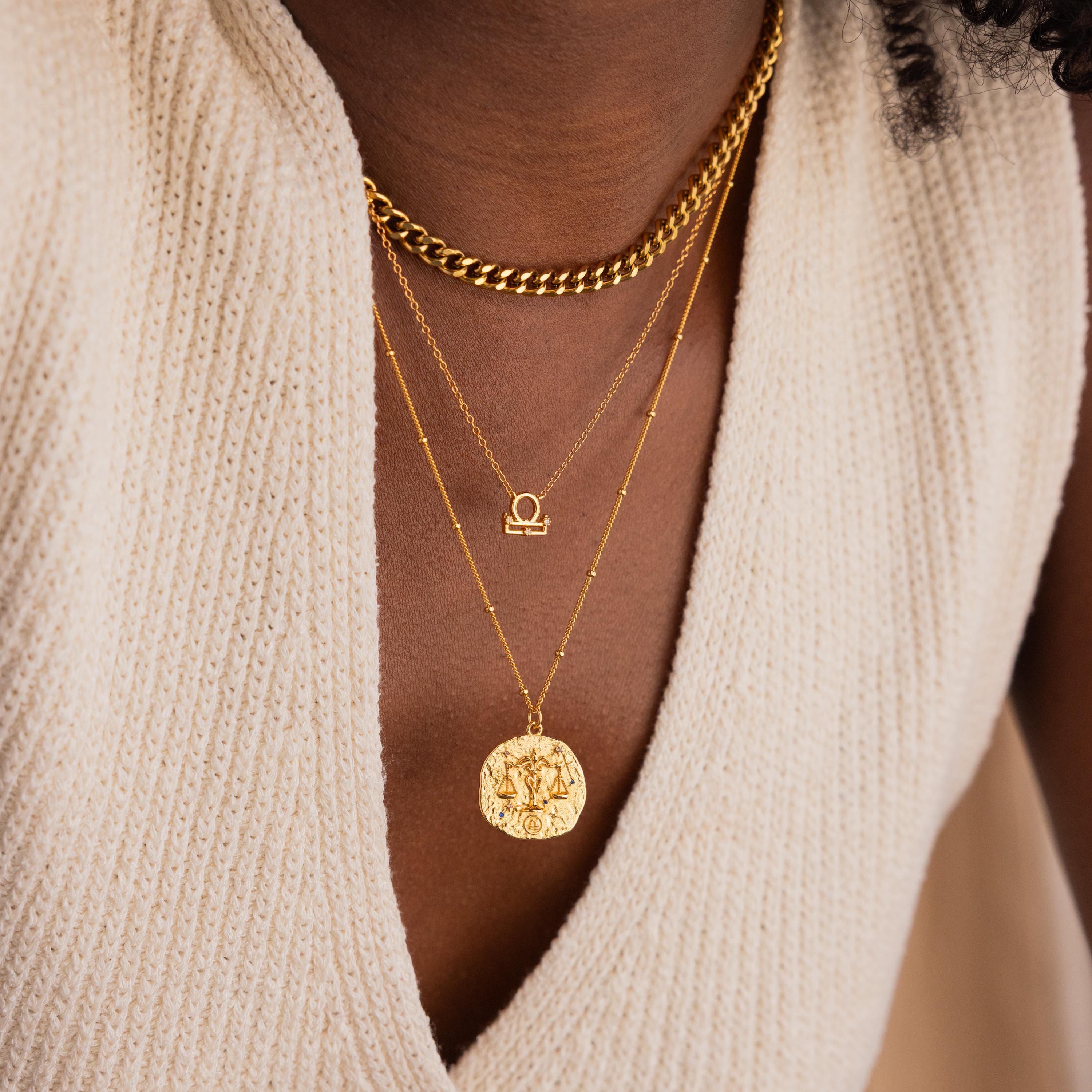 Model wears a gold CZ diamond libra zodiac charm necklace, layered with a thick gold curb chain on top and a hammered libra zodiac circle coin pendant on bottom.