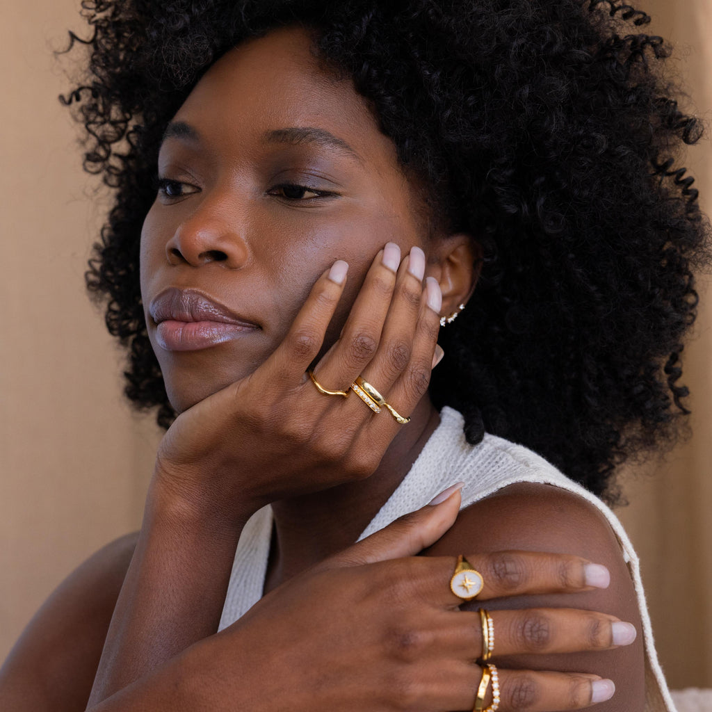 Model's hand reaches up towards her face, while the other one rests on her shoulder, showcasing Celestial Wave Ring alongside other gold rings, including ones such as a Half Dome Ring, Wave Ring, Diamond Beaded Ring, and North Star Signet Ring.