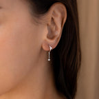 Model wears Silver Star Drop Chain Earrings. The design features a gold hoop earring with a star shaped charm and diamond in the center. A short dangling chain hangs from the hoop with another star shaped charm and diamond at the end.