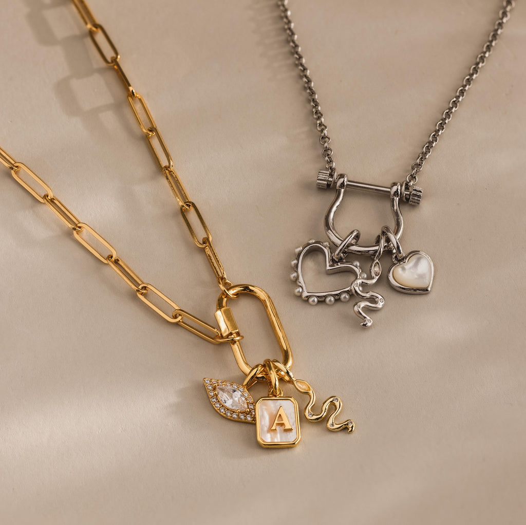 A gold necklace with letter, leaf, and serpent charms, plus a silver necklace featuring a heart and our Viper Snake Charm in Sterling Silver—transform your look with this versatile jewelry duo.