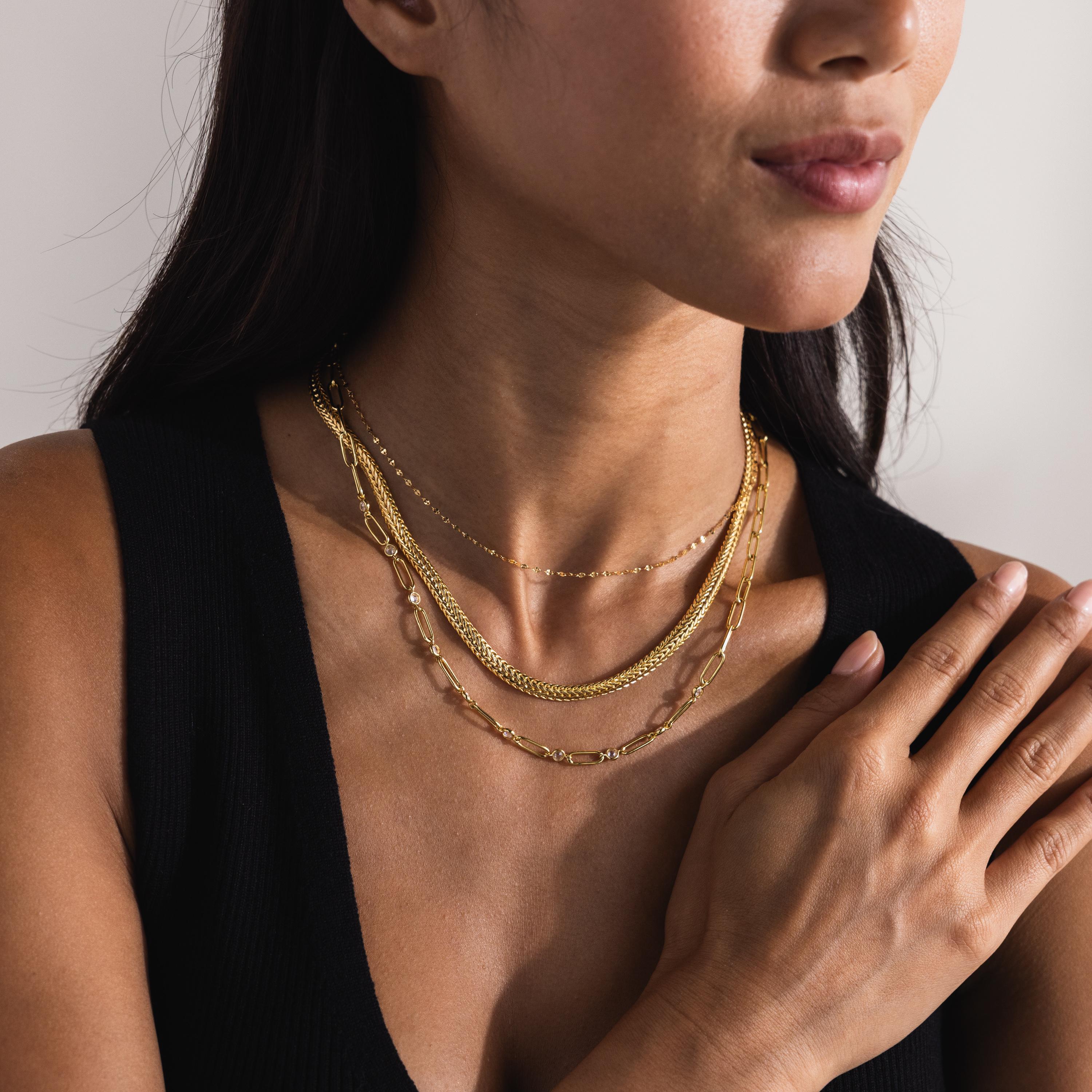 A woman wears a layered necklace stack featuring the gold Foxtail Chain Necklace along with various thinner and thicker gold chains.