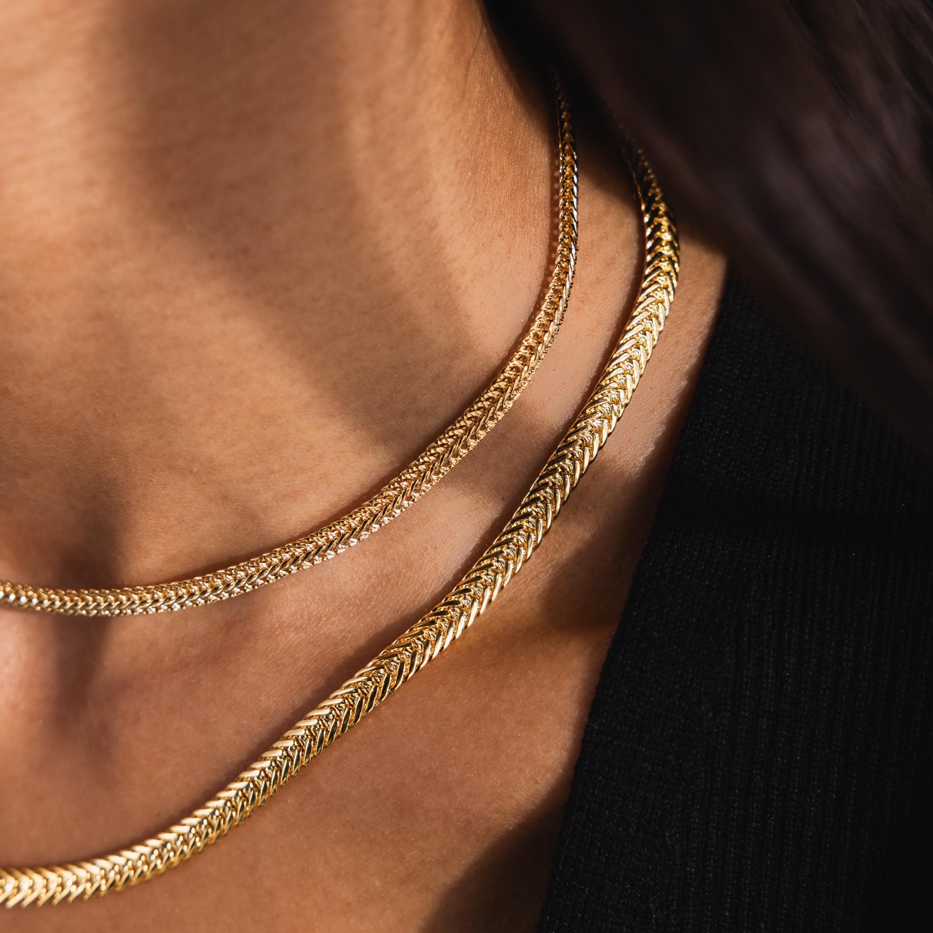Two gold chain necklaces layered on a model’s neck: the Foxtail Chain Necklace on bottom and a Mesh Chain on top.