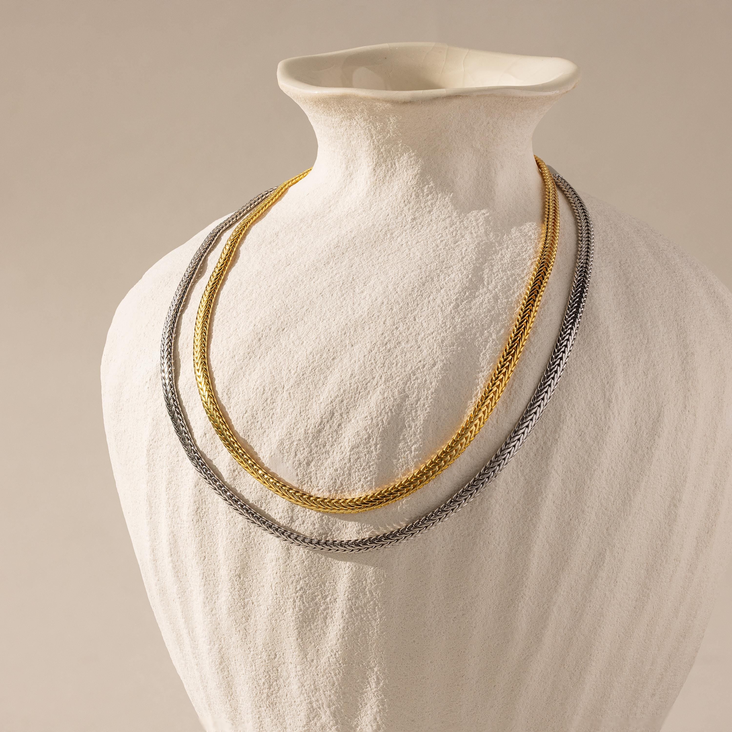 Two Foxtail Chain Necklaces—one in silver and one in 18K gold—layered on a matte white ceramic bust.