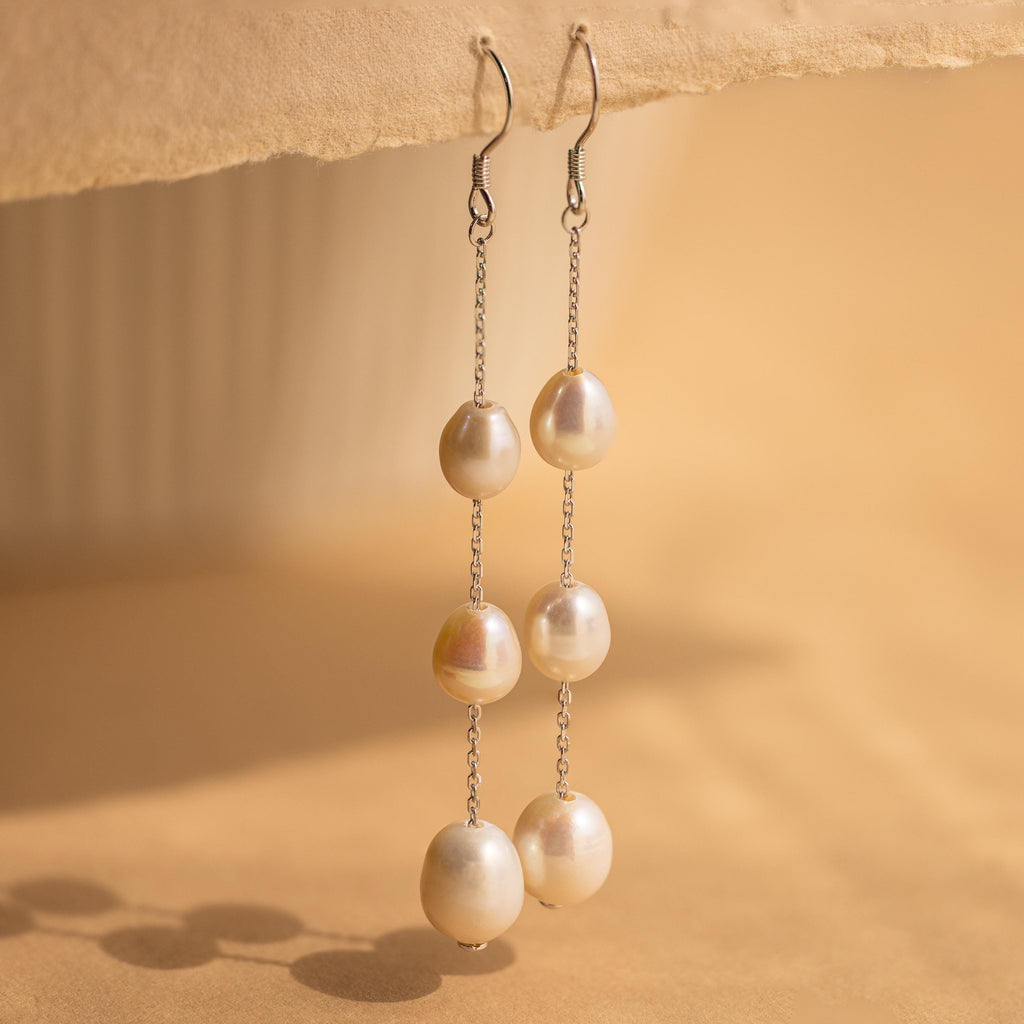 Close-up of a pair of long silver pearl chain earrings hanging from a tan surface.