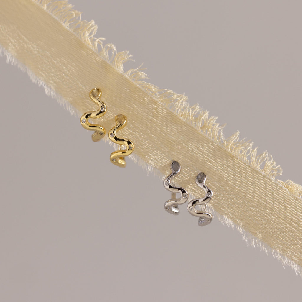 The Pave Viper Huggies, crafted in gold and silver with a sleek serpentine design, are shown on cream-colored fringed fabric.