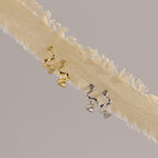 The Pave Viper Huggies, crafted in gold and silver with a sleek serpentine design, are shown on cream-colored fringed fabric.