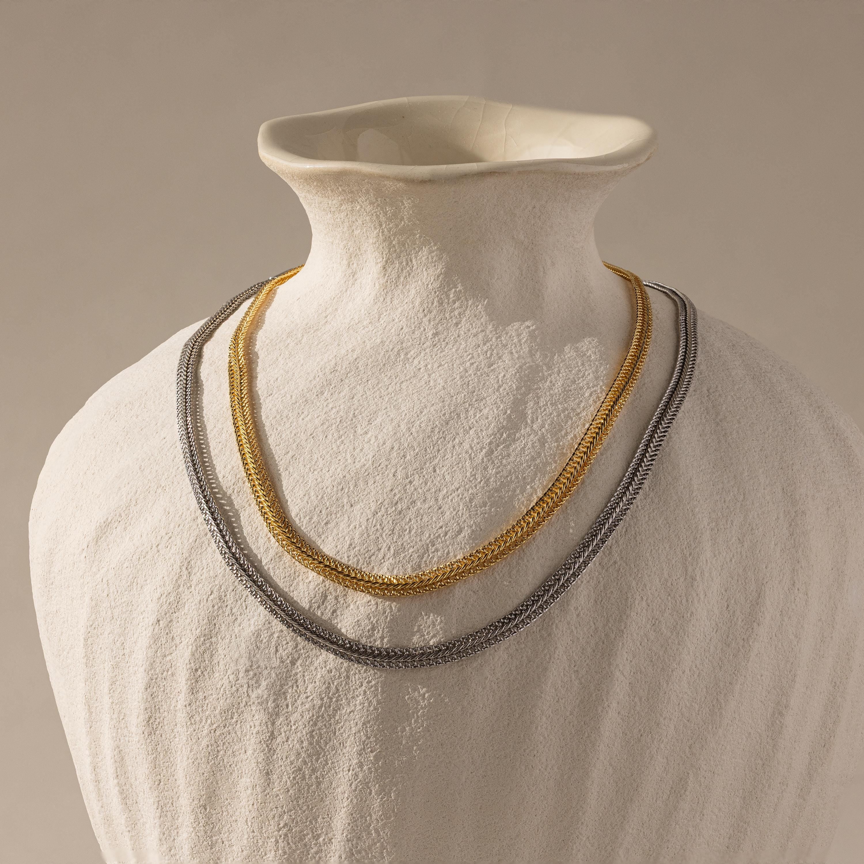 Flat lay of two Thick Mesh Chain Necklaces displayed on a sculptural ceramic bust—one in gold and the other in silver