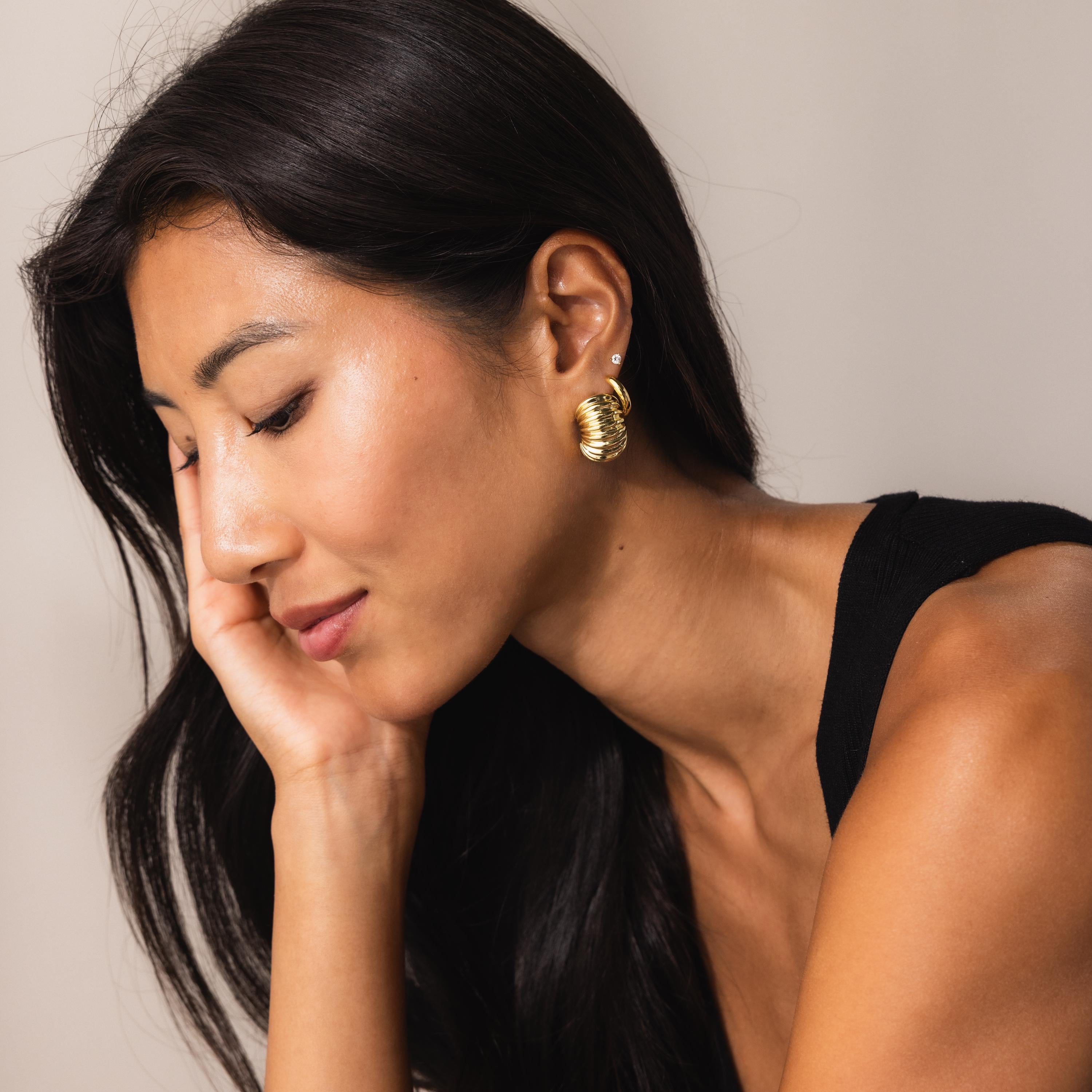 Model in profile pose wearing chunky gold ribbed earrings with oval hoop and diamond stud; the earrings catch the light and complete her minimal, elegant look.
