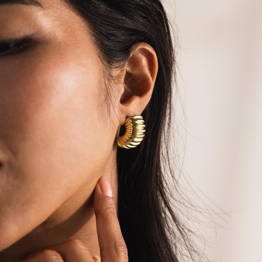 A close-up view of a woman's ear wearing a chunky ribbed huggie earring in 18K gold; the design features deep ridges along the curved, dome-like shape that hugs the earlobe snugly, with high polish catching the light for a lustrous finish