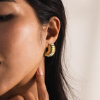 A close-up view of a woman's ear wearing a chunky ribbed huggie earring in 18K gold; the design features deep ridges along the curved, dome-like shape that hugs the earlobe snugly, with high polish catching the light for a lustrous finish