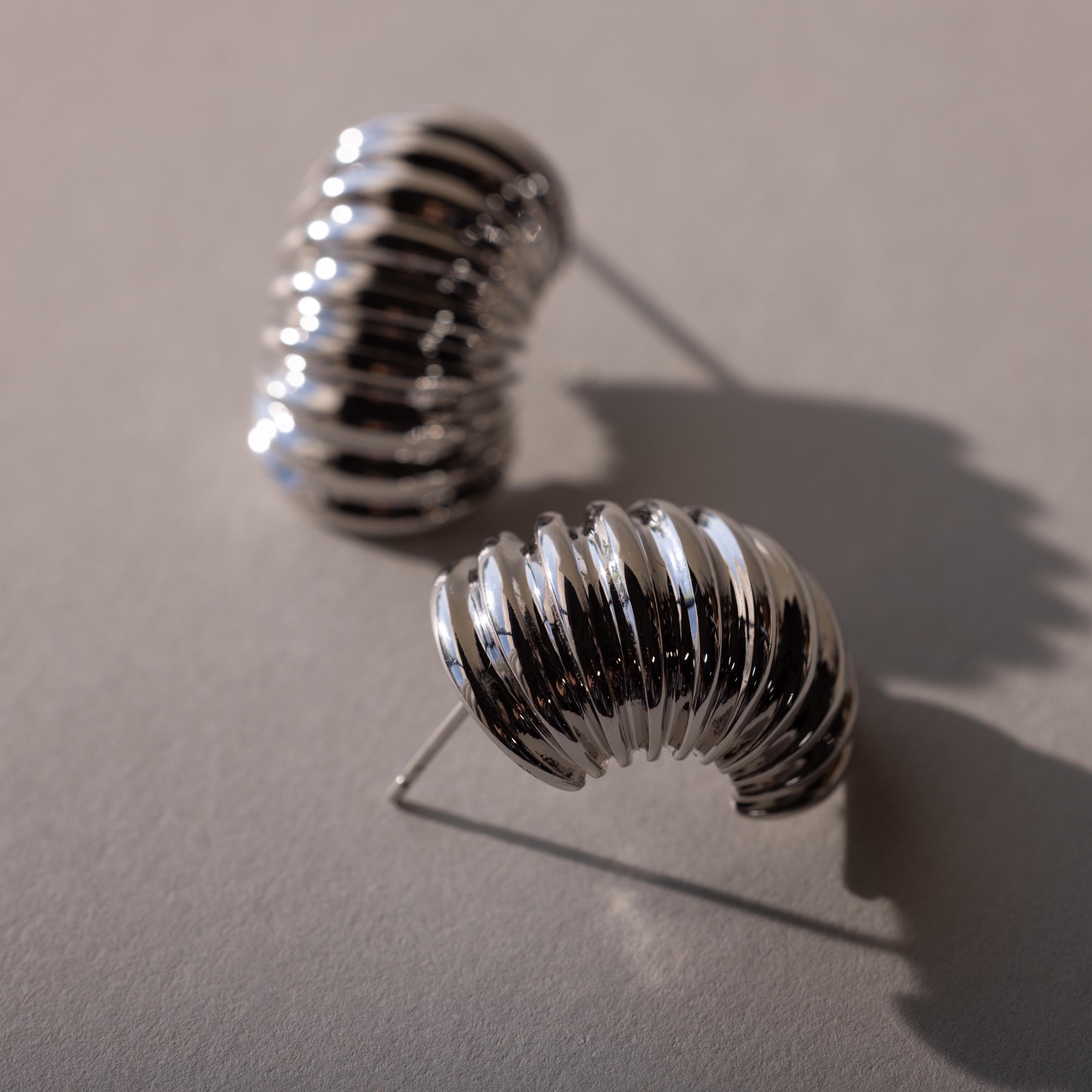 The Chunky Ribbed Hoops are two shiny, ribbed silver hoops with curved, chunky designs—perfect statement earrings for your ear stack, shown on a neutral background.