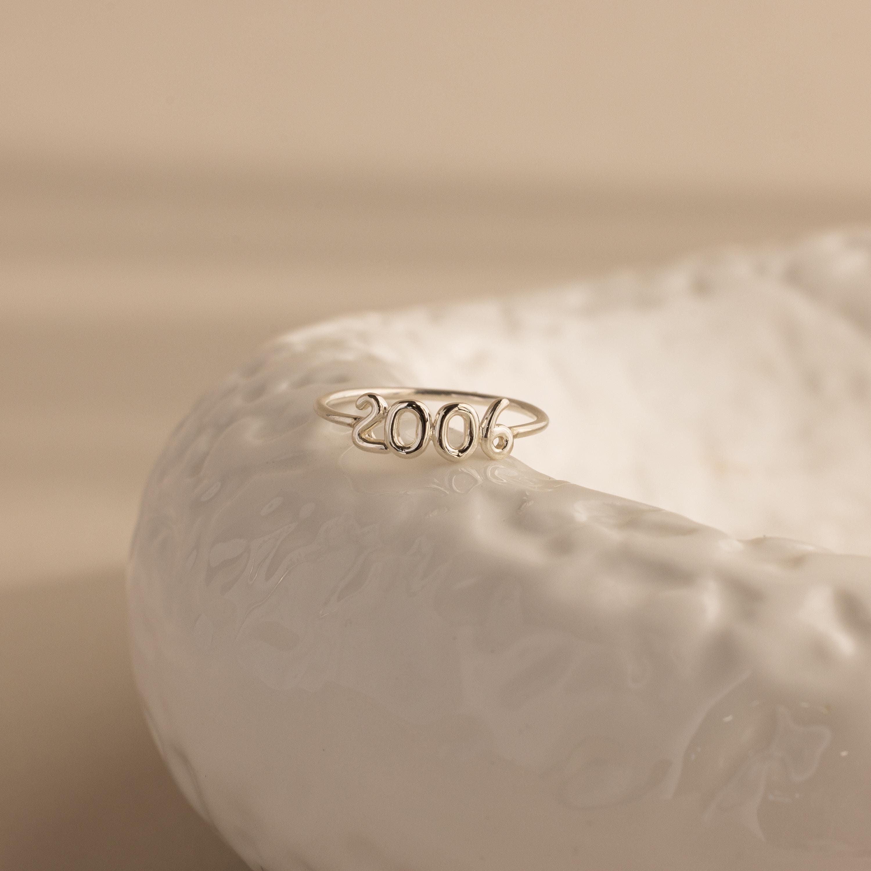 A silver “2006” Bubble Numbers Ring is perched on a textured white ceramic dish.