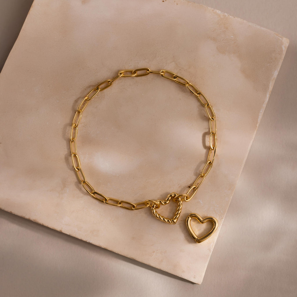 Heart Carabiner Paperclip Bracelet in gold laid flat with removable heart charm on a beige stone surface.