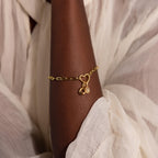 Close-up of the Heart Carabiner Paperclip Bracelet in gold, worn on the wrist of a dark-skinned model. The bracelet is composed of elongated, rectangular paperclip-style links with a polished finish and a twisted heart carabiner in the middle.