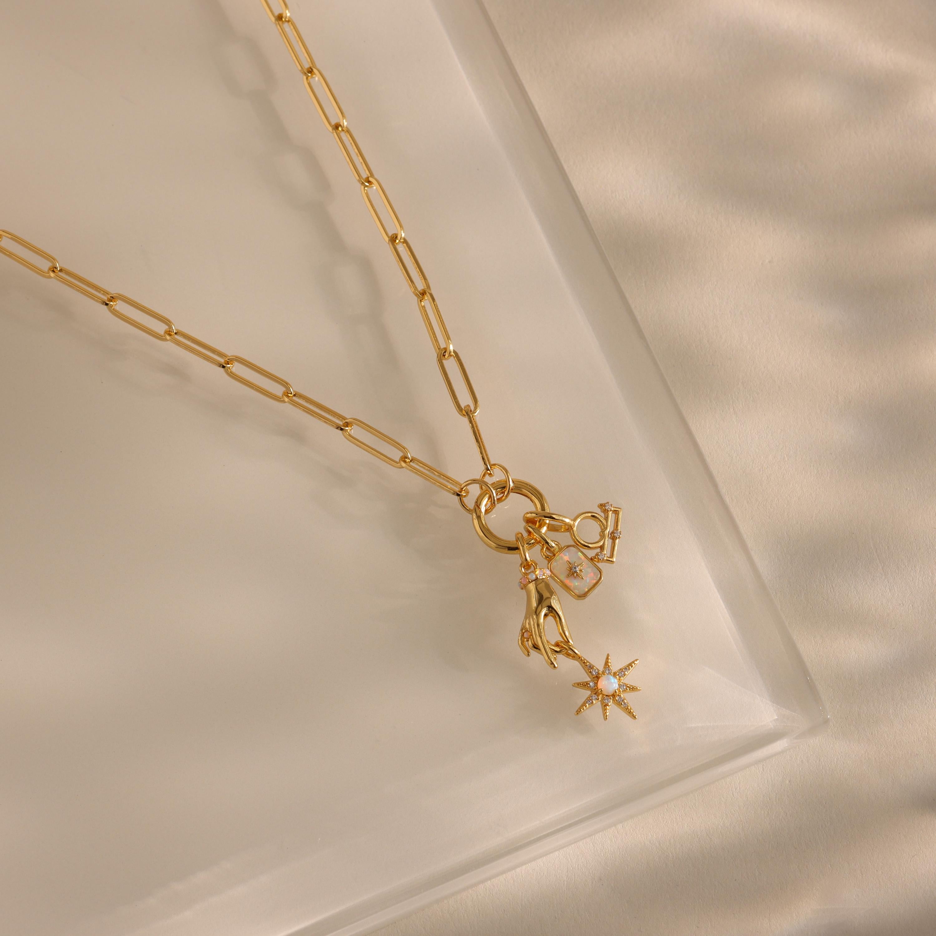 A gold paperclip chain necklace with multiple charms—zodiac symbols, a pearl square, and a star—is laid flat on a clear surface.