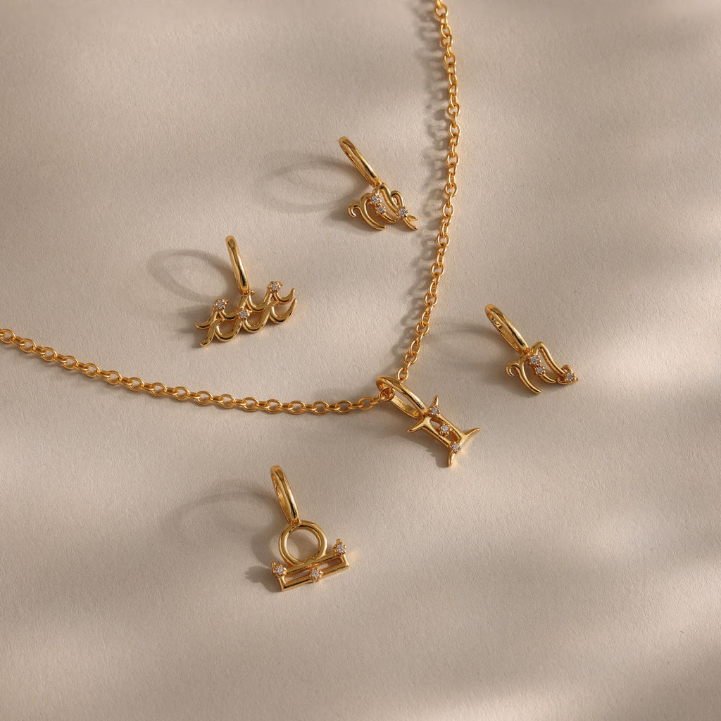 A flat-lay of gold zodiac charm pendants and a matching chain on a soft beige surface, featuring finely crafted zodiac symbols such as Aquarius, Libra, Gemini, Pisces, and others, each adorned with delicate cubic zirconia accents for subtle sparkle.