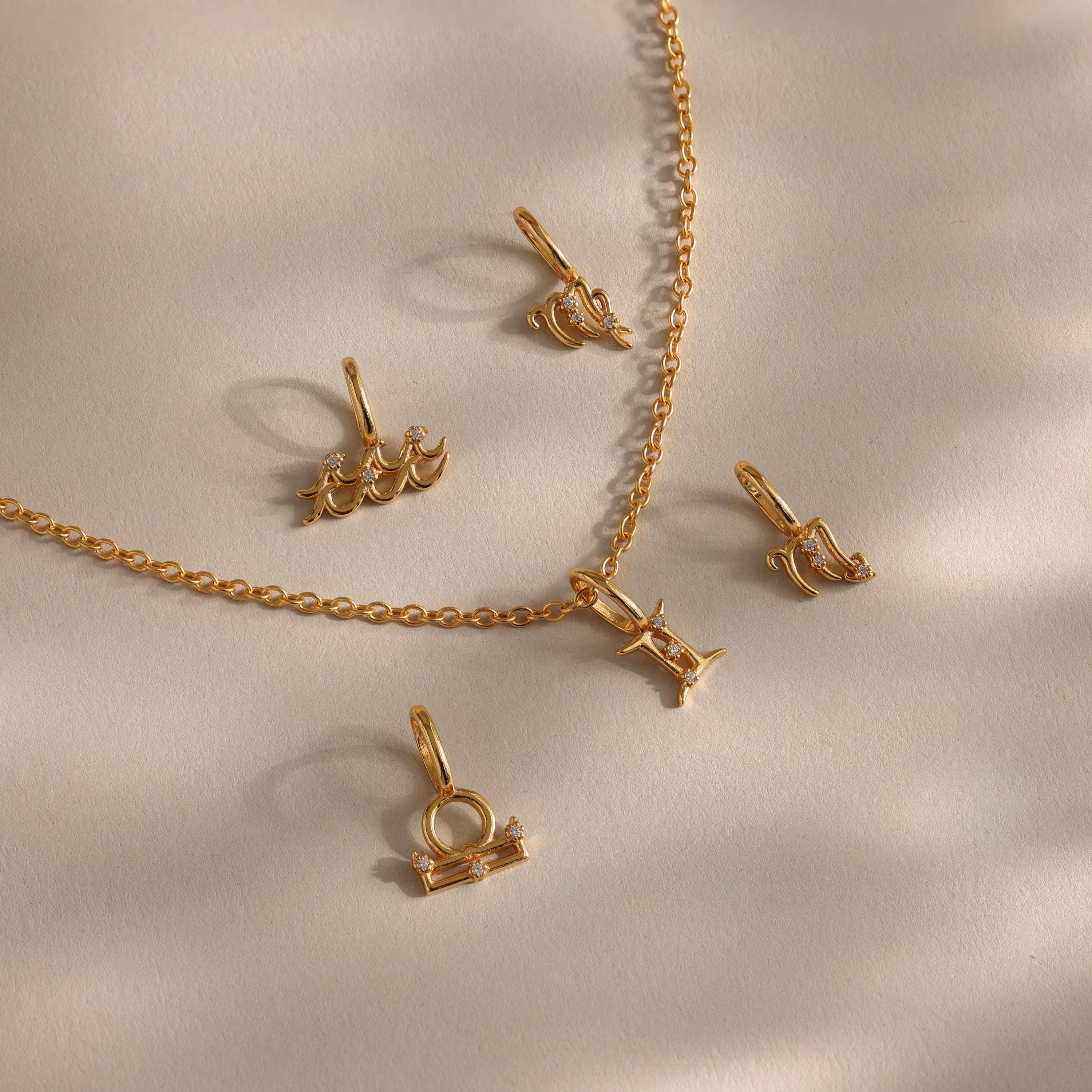 A flat-lay of gold zodiac charm pendants and a matching chain on a soft beige surface, featuring finely crafted zodiac symbols such as Aquarius, Libra, Gemini, Pisces, and others, each adorned with delicate cubic zirconia accents for subtle sparkle.