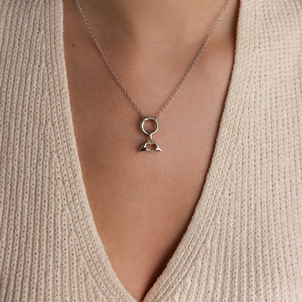 A silver Heart Hands charm is worn on a necklace with a round connector and fine silver chain over a beige knit top.