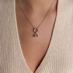 A silver Heart Hands charm is worn on a necklace with a round connector and fine silver chain over a beige knit top.