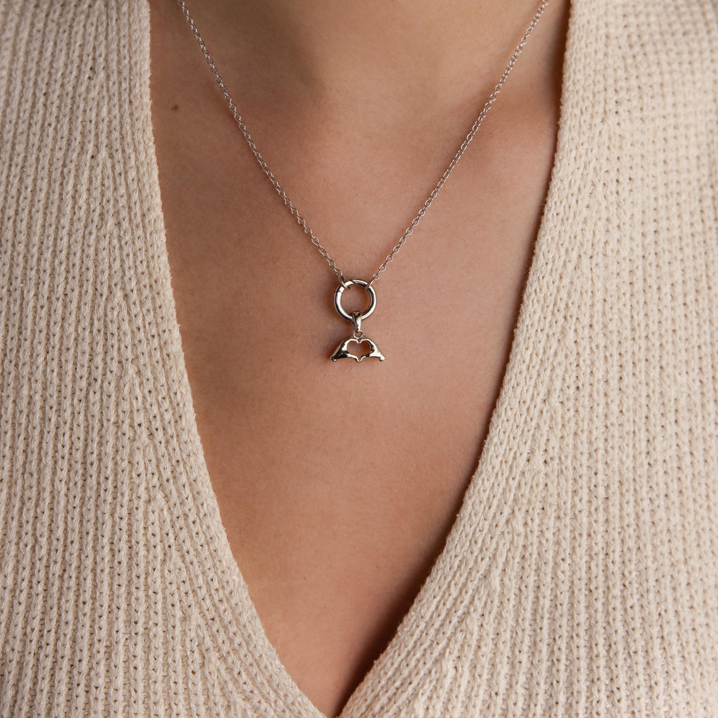 A silver Heart Hands charm is worn on a necklace with a round connector and fine silver chain over a beige knit top.