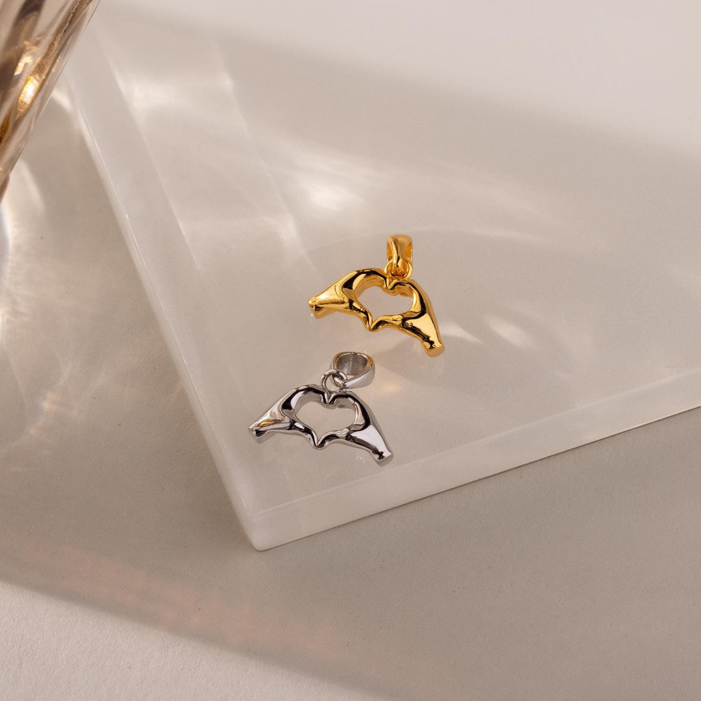 Gold and silver Heart Hands charms lie side-by-side on a glossy cream-toned display platform, catching soft, natural light.