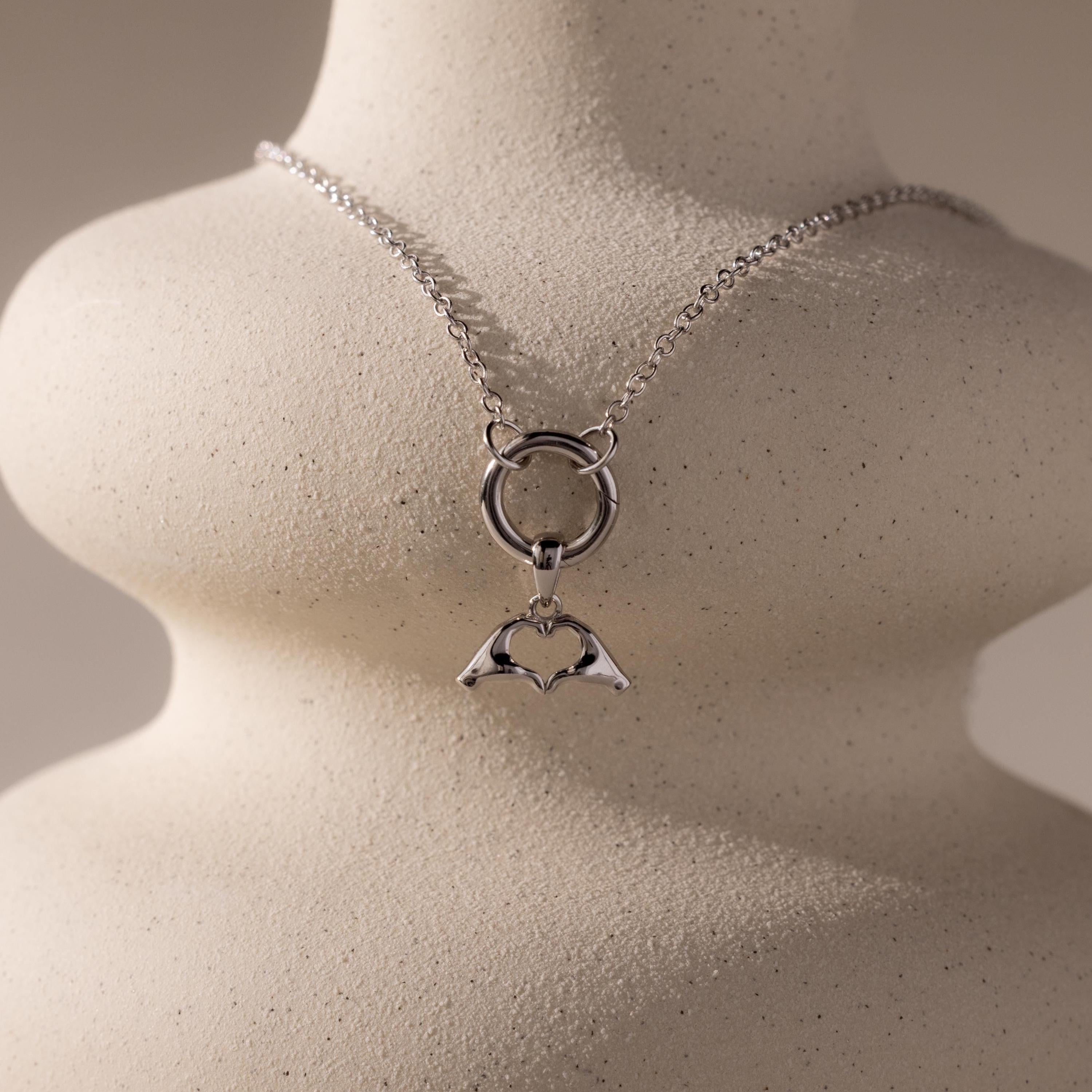 The silver Heart Hands charm is styled on a sculptural off-white display with a matching silver chain and round connector.