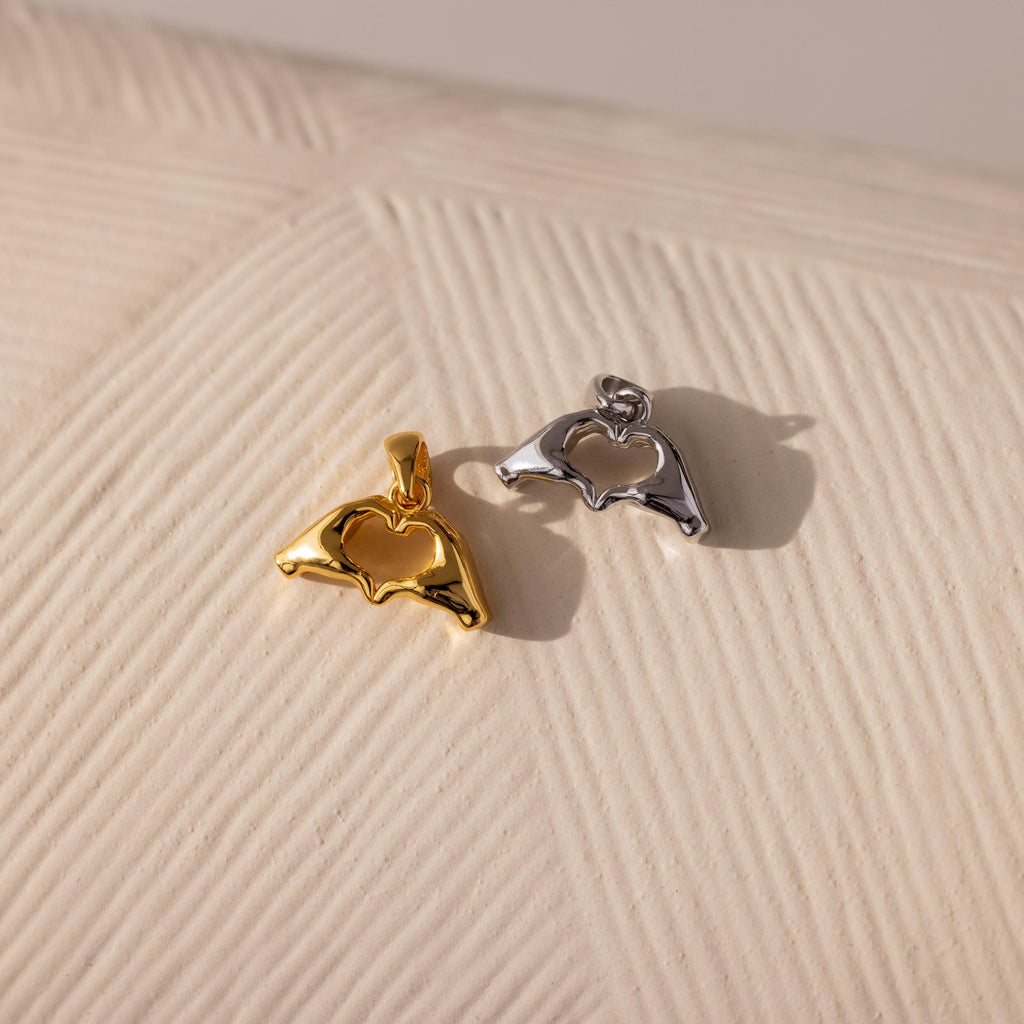 Two metal Heart Hands charms—one in gold and one in silver—are displayed on a textured cream surface, each shaped like two hands forming a heart with a cut-out center.