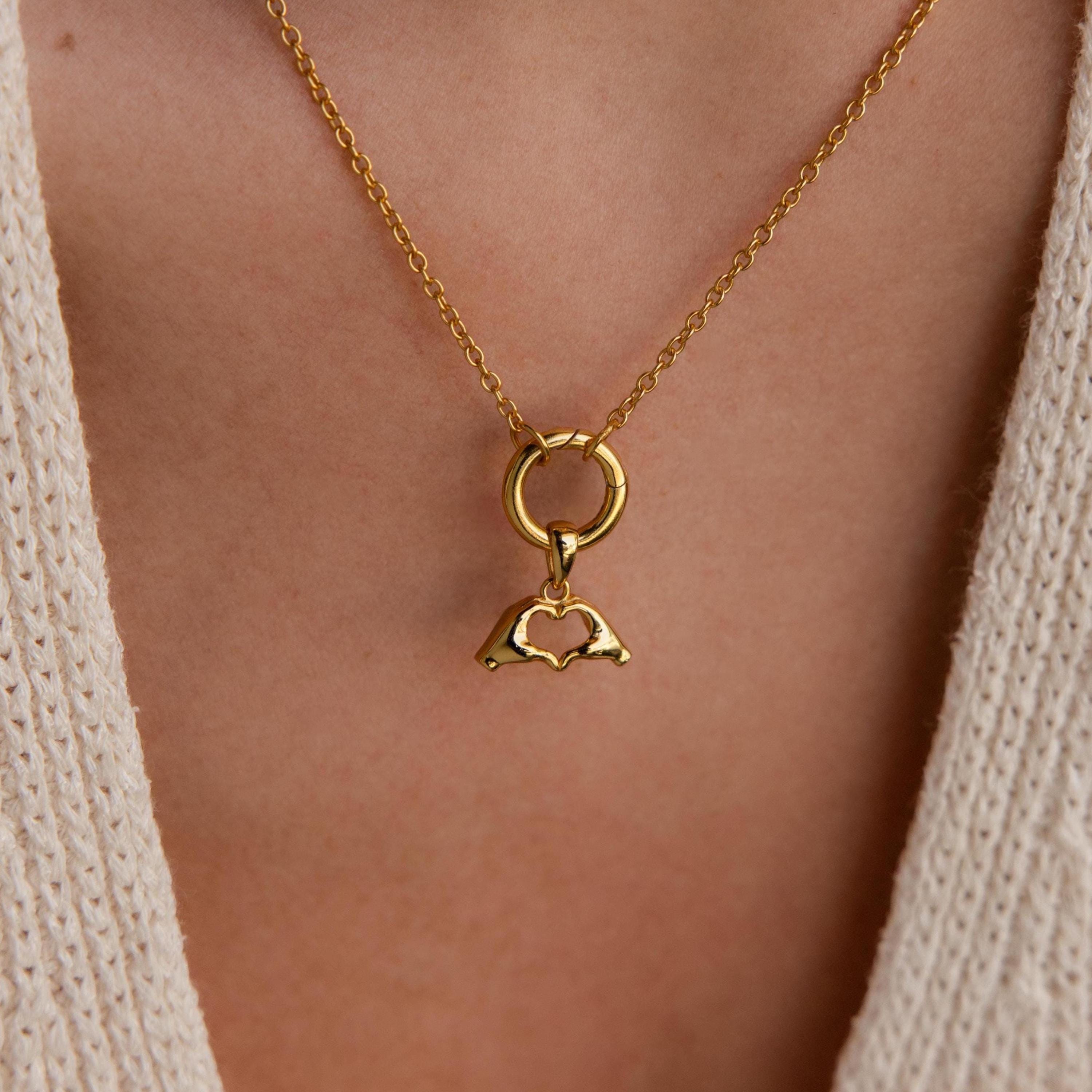 A gold Heart Hands charm hangs from a circular bail on a gold chain necklace worn over a cream knit top.