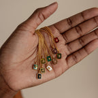A hand holds several gold necklaces with colorful rectangular gemstone pendants, including the North Star Garnet Necklace and a radiant crystal pendant.