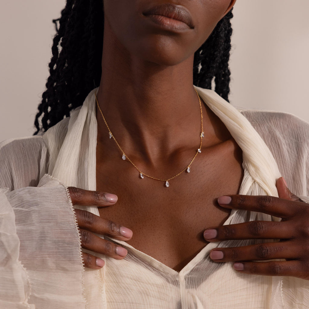 Model wearing the gold necklace with both hands gently framing it over a cream blouse.