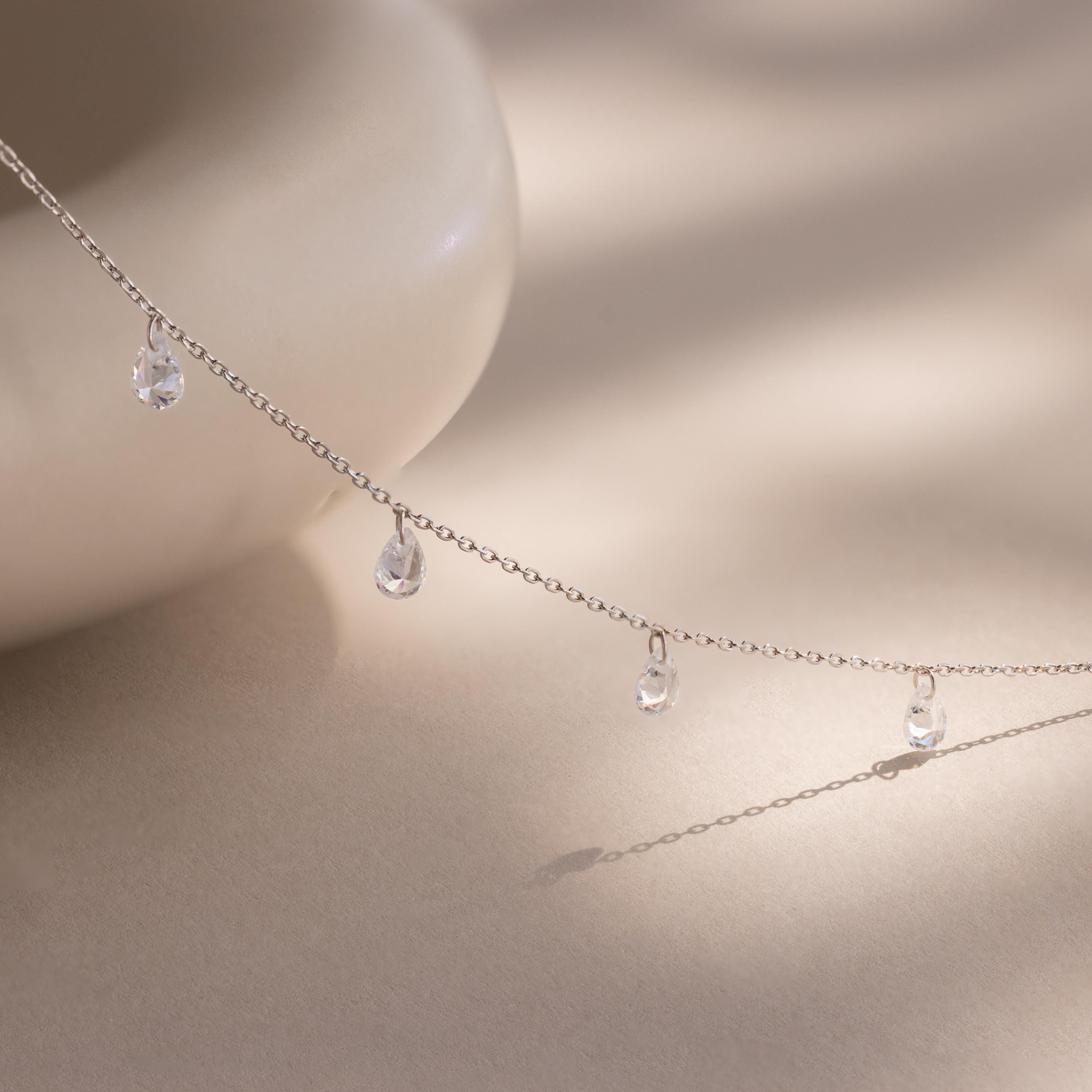 Elegant silver chain necklace featuring evenly spaced teardrop-shaped diamond charms, delicately draped over a textured cream-colored cylindrical display.