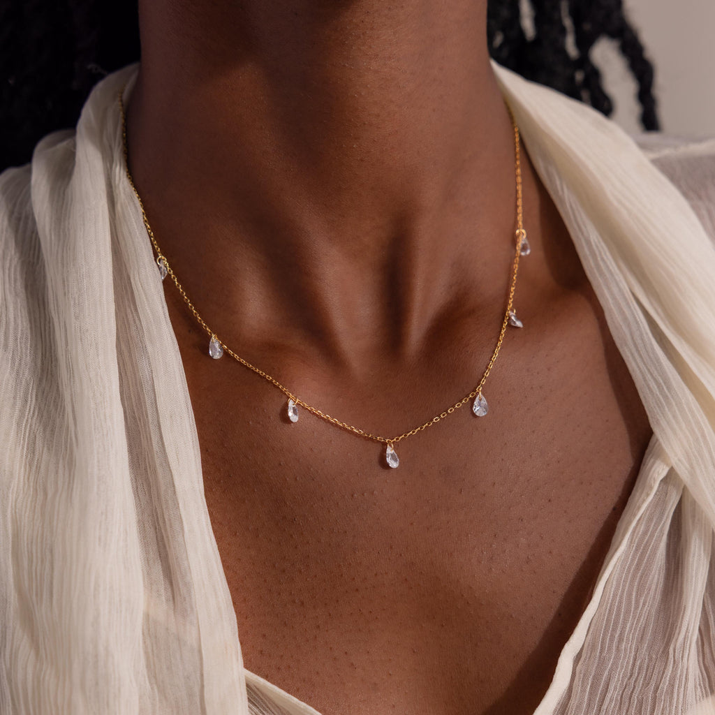 A close-up view of a woman wearing a delicate gold chain necklace with small, teardrop-shaped diamond-like pendants evenly spaced along the chain.