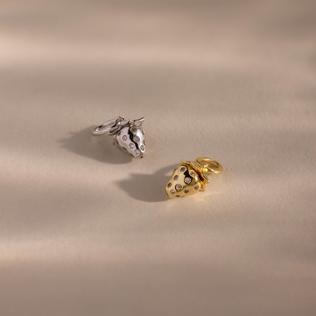 Gold and silver strawberry charms are placed on a warm beige background, both detailed with scattered inset crystals.