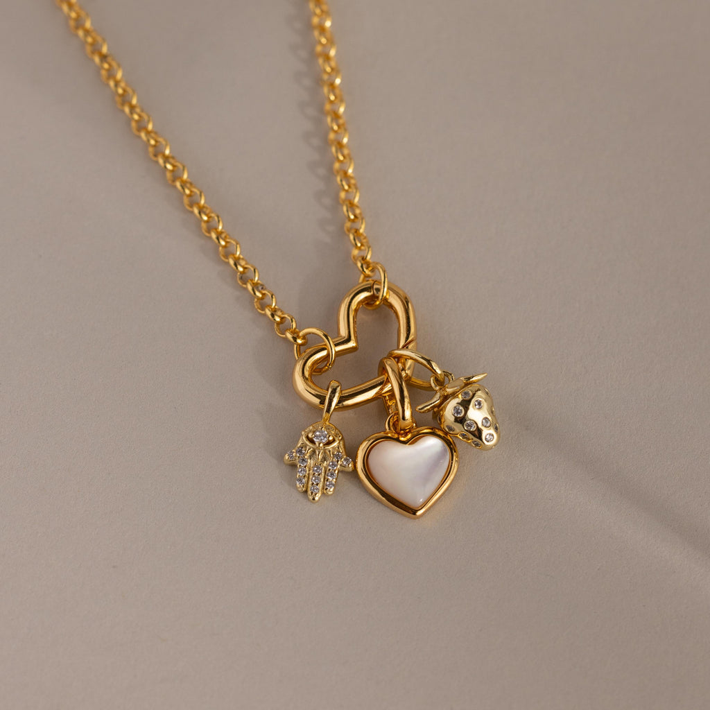 A gold chain necklace with a heart-shaped connector holds three charms: a pavé hamsa, a mother-of-pearl heart, and a strawberry charm, all resting on a soft matte surface.