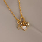 A gold chain necklace with a heart-shaped connector holds three charms: a pavé hamsa, a mother-of-pearl heart, and a strawberry charm, all resting on a soft matte surface.