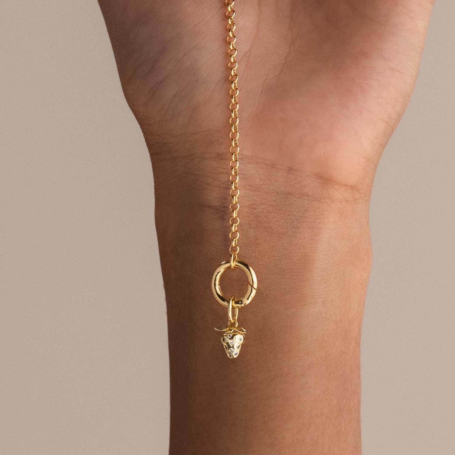A delicate gold rolo chain bracelet is held vertically, featuring a single dangling gold strawberry charm with sparkling crystal details.