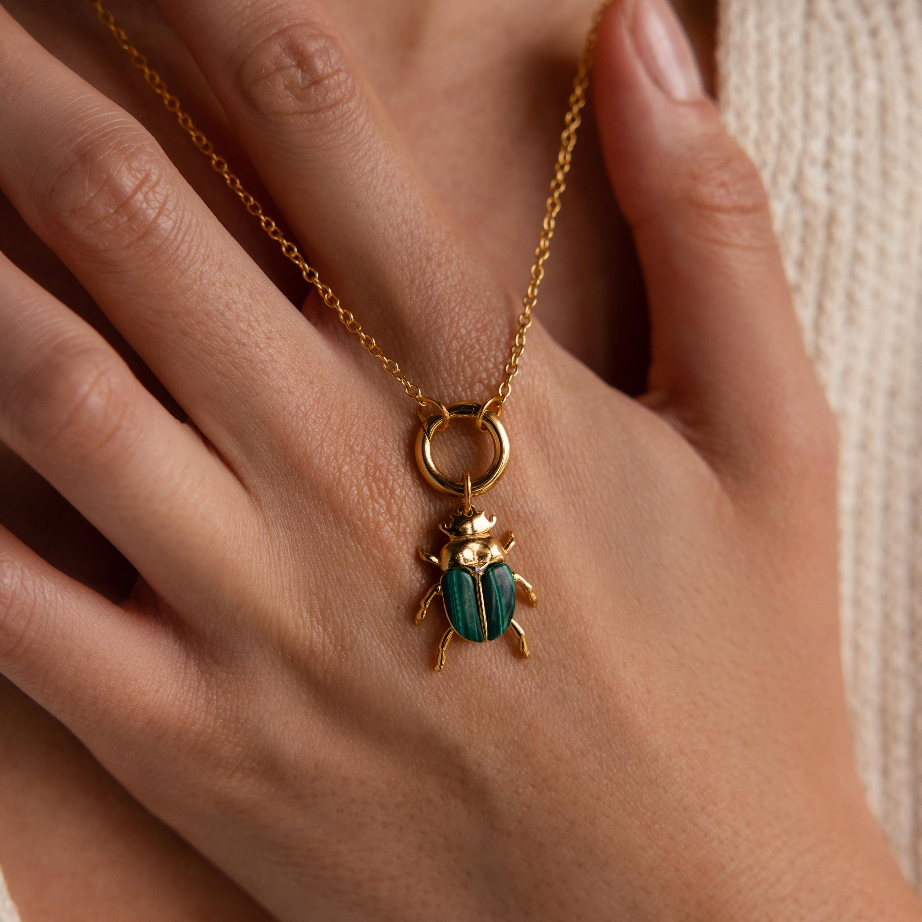 A close-up of a hand gently resting over a gold chain necklace featuring a gold and green malachite beetle charm with a crown motif on its head. The charm is suspended from a circular Rolo clasp.