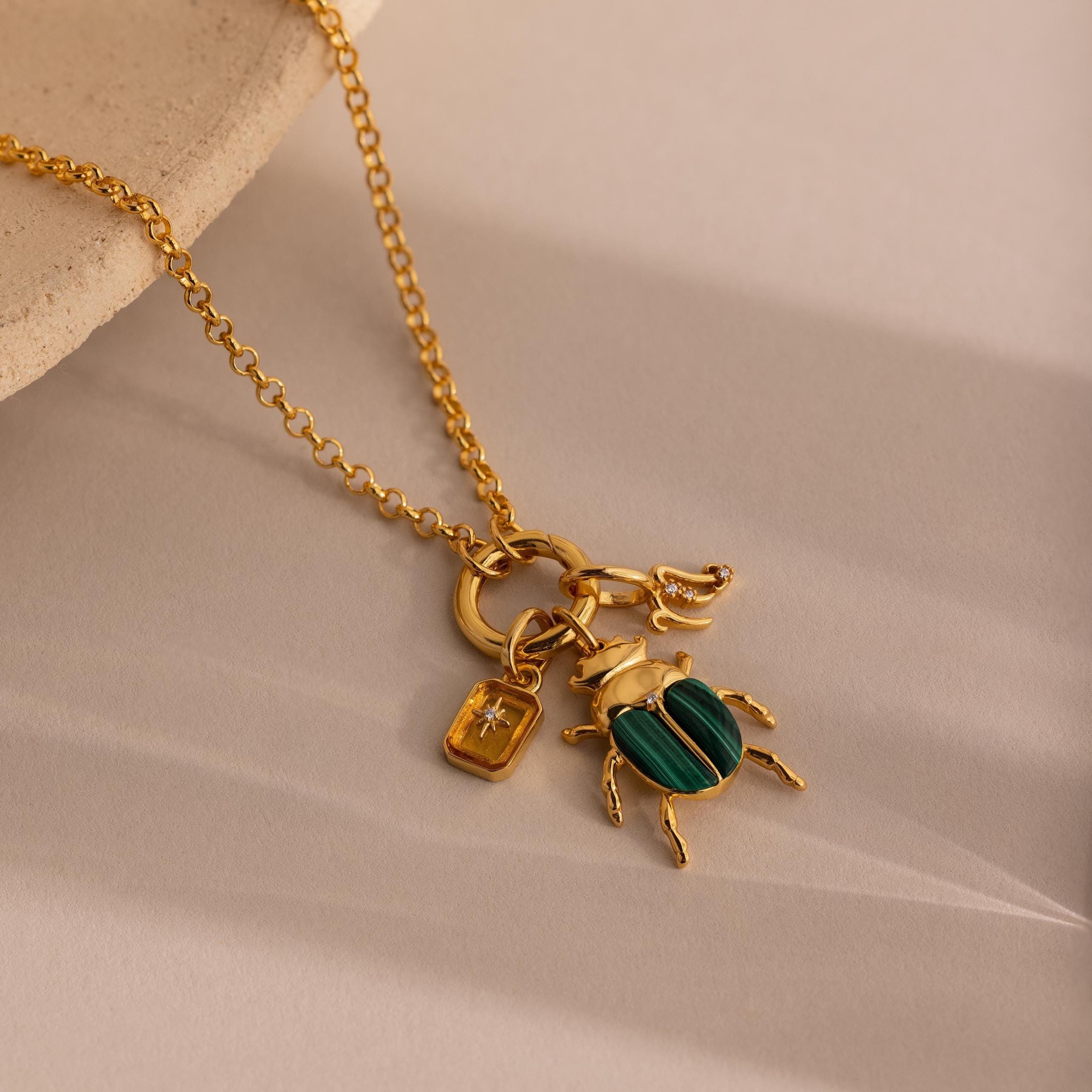 The Malachite Beetle Charm is shown styled with a gold necklace on a light stone surface. Other pendants  including a rectangular North Star Birthstone Charm and a Zodiac Charm