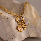 The Opal Sun Charm, opal disc charm, and an “S” initial charm are shown hanging from a round clasp set atop a rough-edged beige stone, with a gold paperclip chain looped around