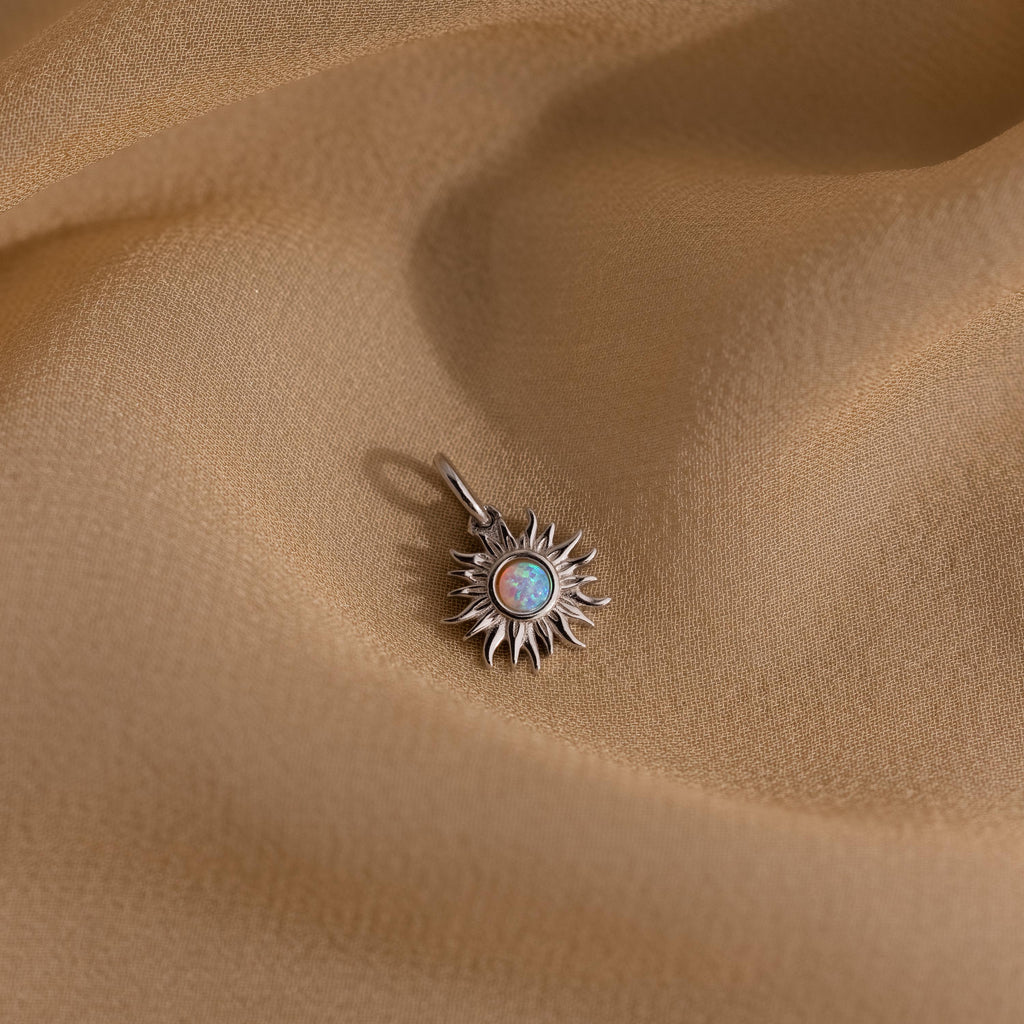 A silver version of the Opal Sun Charm placed on flowing golden fabric, showcasing the opalescent center stone and fine sunray details.