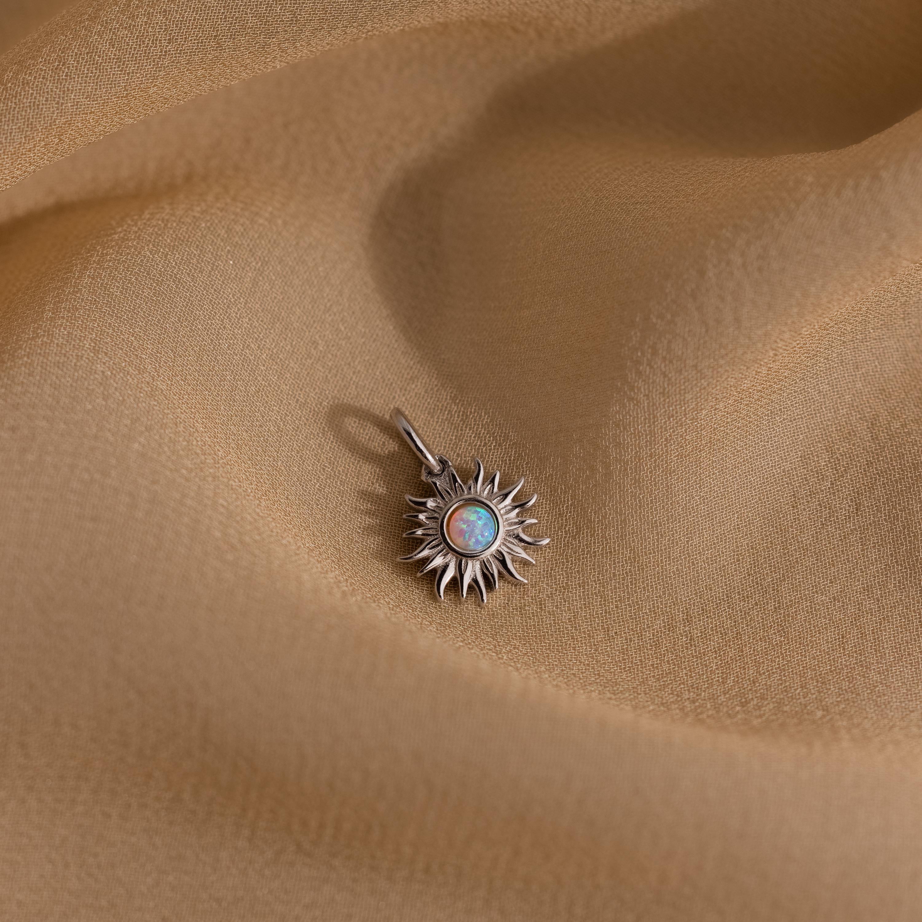 A silver version of the Opal Sun Charm placed on flowing golden fabric, showcasing the opalescent center stone and fine sunray details.