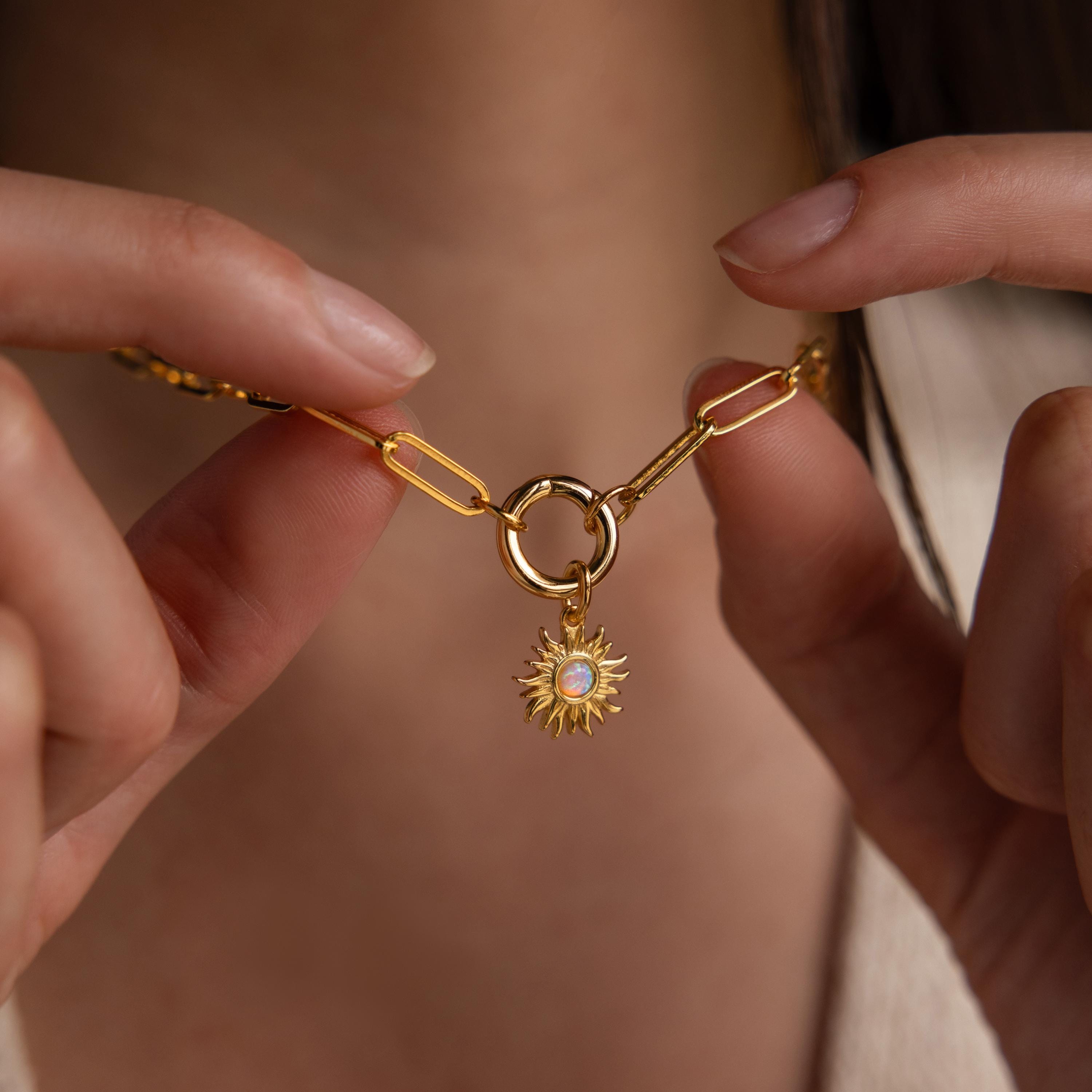 A person holds a gold paperclip chain necklace with a circular clasp, from which the Opal Sun Charm dangles prominently at the center.